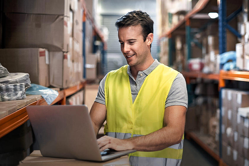 This image shows a man wearing a yellow vest as he stands in a shipping warehouse. He is using the intelligent document processing software on his laptop to automate unstructured data.