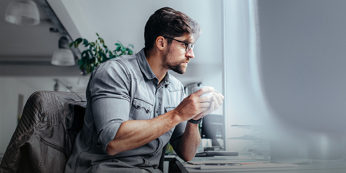 The image shows a man wearing a gray shirt sitting at a desk, looking at something on his computer screen. He is wearing glasses and has his hands around a coffee mug. It looks like he is pondering the What is Robotic Process Automation Software and How it Works. There is a window behind him with tree in the background. The image is taken from a low angle, looking up at the man from behind his desk.